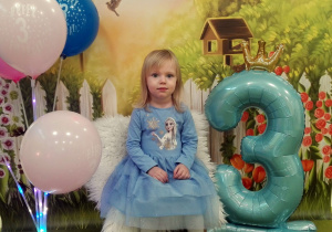 Ala siedzi na urodzinowym krześle. Obok niej stoi cyfra trzy oraz kolorowe balony.