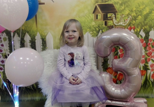 Hania siedzi na urodzinowym krześle. Obok niej stoi cyfra trzy oraz kolorowe balony.