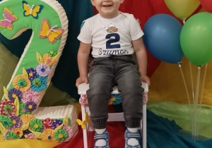 Szymon siedzi na urodzinowym krześle. Obok niego stoi styropianowa liczba dwa oraz kolorowe balony.