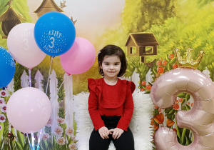 Roma siedzi na urodzinowym krześle. Obok stoi cyfra trzy i kolorowe balony.