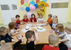 Dzieci siedzą przy stole i jedzą urodzinowe smakołyki.