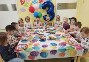 Dzieci siedzą przy stole i jedzą urodzinowe smakołyki.