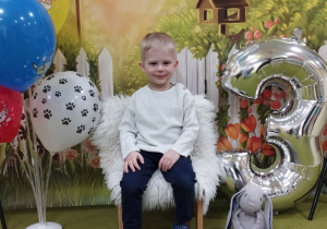 Kacper siedzi na urodzinowym krześle, obok stoją balony i cyfra trzy.