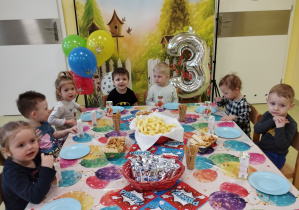 Dzieci siedzą przy urodzinowym stole i jedzą smakołyki.