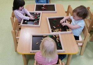 Dzieci siedzą przy stolikach i bawią się tackami sensorycznymi z kolorowym makaronem.