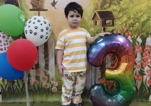 Adam stoi przy cyfrze trzy. Obok stoją kolorowe balony.