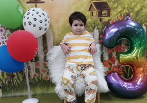 Adam siedzi na urodzinowym krześle. Obok stoi cyfra trzy i kolorowe balony.