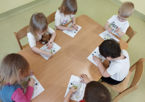 Dzieci siedzą przy stoliku i kredkami kolorują żabkę.