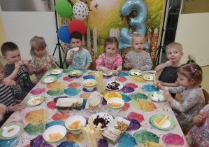 Dzieci siedzą przy urodzinowym stole i jedzą smakołyki.
