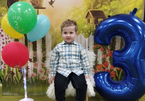 Olek siedzi na urodzinowym krześle. Obok stoi cyfra trzy i kolorowe balony.