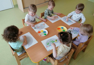 Dzieci siedzą przy stolikach i kolorują tęcze.
