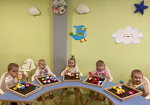 Dzieci siedzą przy stole i bawią się kolorowymi pomponami.