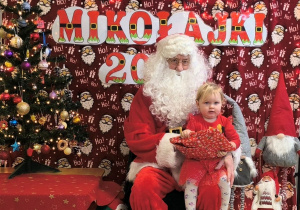 Mikołaj siedzi na krześle i pozuje do zdjęcia. Na kolanach Mikołaja siedzi Amelka i trzyma w rękach prezent mikołajkowy.