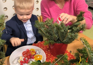 Tobiasz oraz jego mama siedzą przy stole i wspólnie ozdabiają świąteczny stroik.