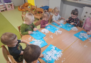 Dzieci siedzą przy stoliku i wykonują pracę plastyczną. Za pomocą małych białych ścinków wyklejają bałwana.