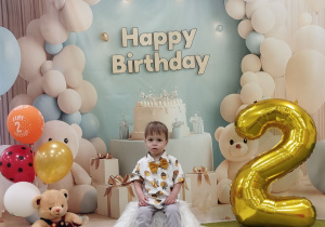 Adaś pozuje do pamiątkowego zdjęcia, obok stoją balony i cyfra dwa.
