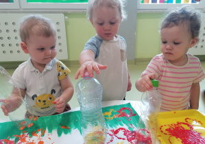 Jakub, Maksymilian i Emilia przy pomocy plastikowych butelek stemplują obrazek przedstawiający łąkę.