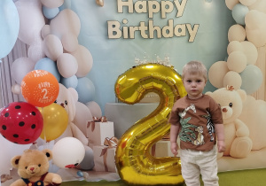 Ignacy pozuje do pamiątkowego zdjęcia, obok stoją balony i cyfra dwa.