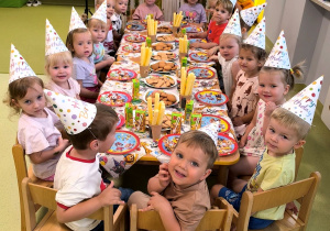 Dzieci pozują do zdjęcia siedząc przy stolikach podczas przyjęcia urodzinowego.
