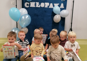 Chłopcy siedzą na dywanie i pozują z upominkami z okazji Dnia Chłopca.
