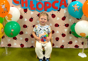 Ignacy siedzi na krzesełku. Obok stoją kolorowe balony. zdjęcie zrobione jest na białym tle z czerwonymi biedronkami.