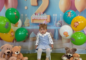 Tola siedzi na krzesełku. Za nią wisi urodzinowe tło z cyfrą dwa. Obok stoją kolorowe balony i siedzą misie.