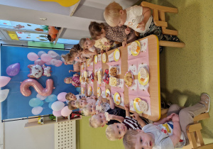 Dzieci siedzą przy stole i jedzą urodzinowy poczęstunek.