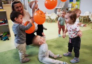 Dzieci bawią się pomarańczowymi balonami łapiąc je w ręce, podrzucając.