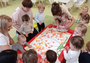Dzieci przyklejają pomarańczowe kartki do konturu dyni na dużym brystolu.