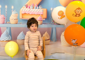 Lavrentii siedzi na małym krzesełku na tle urodzinowym z cyfrą numer dwa. Obok stoją balony na stojaku.