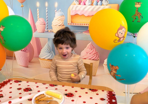 Lavrentii siedzi przy stoliku na tle urodzinowym z cyfrą dwa i balonami. na stole stoją słodkie przekąski.