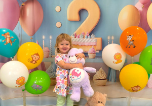 Antosia stoi na tle urodzinowym z cyfrą dwa. W rękach trzyma misia a drugi miś siedzi obok na podłodze. Obok stoją kolorowe balony.