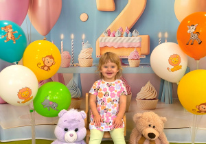 Antosia siedzi na krzesełku. Obok z dwóch stron siedzą misie i stoją kolorowe balony na stojakach.