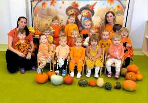 Zdjęcie grupowe. Dzieci i opiekunki siedzą i stoją na dywanie na tle z dyniami. Na podłodze przed dziećmi leżą ułożone dynie.