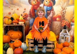 Tosia siedzi na skrzynce i pozuje do zdjęcia. Obok po dwóch stronach leżą dynie.