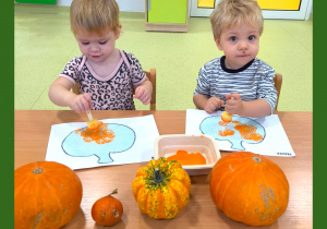 Dwójka dzieci siedzi przy stole i maluje za pomocą gąbki dynie wycięte z folii bąbelkowej i naklejone na kartkę.