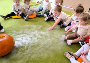 Dzieci siedzą na dywanie. Na środku stoi duża dynia która ma wycięte oczy i buzię. Dzieci oglądają doświadczenie z użyciem suchego lodu.