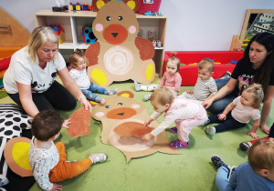 Dzieci z pomocą opiekunki układają misia z pociętych elementów.