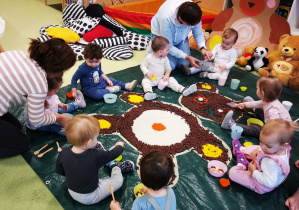 Dzieci z opiekunkami siedzą na macie i bawią się w przesypywanie sypkich artykułów ułożonych w postaci misia.