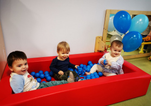 Dzieci siedzą w małym basenie, bawią się niebieskimi piłeczkami.