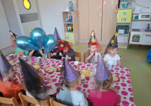 Dzieci siedzą na poczęstunku z okazji Andrzejek. Na głowach mają kolorowe czapeczki.