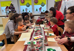Dzieci wraz z rodzicami wykonują kartkę bożonarodzeniową przy stolikach.