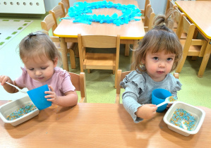 Dziewczynki siedzą przy stoliku i przesypują niebieski ryż z pojemnika do niebieskiego kubeczka.