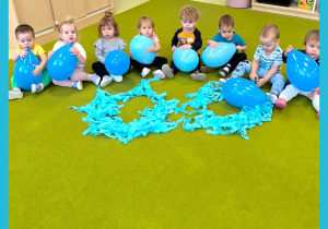 Dzieci siedzą na dywanie. Każde trzyma w ręku niebieski balon. Przed dziećmi leży hula hop udekorowany niebieską bibułą.