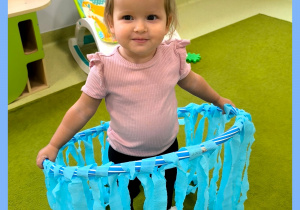 Amelka stoli na dywanie i trzyma hula hop udekorowany niebieską bibułą.