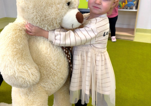 Apolonia stoi na dywanie i przytula dużego pluszowego misia.