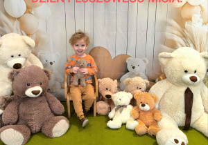 Filip siedzi na krześle. Obok stoją pluszowe misie. Za Filipem powieszone jest tło z okazji pluszowego misia.