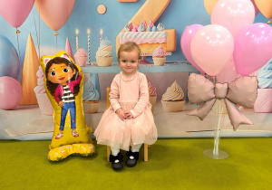 Apolonia siedzi na krzesełku. Obok stoją balony. Za Apolonią powieszone jest urodzinowe tło.