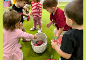 Na dywanie postawiony jest biały wiklinowy koszyk. Dzieci zbierają papierowe czerwone jabłuszka i wrzucają je do koszyka.