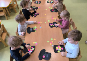 Dzieci siedzą przy stolikach i za pomocą kolorowych gwiazdek wyciętych z papieru wyklejają cylinder.